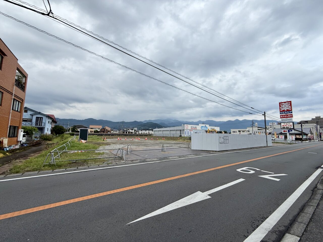 道路ぞいから見たコスモス長松寺店の建設地。奥にウエルシア甲府池田店と山並みが見える(2026年7月18日撮影)